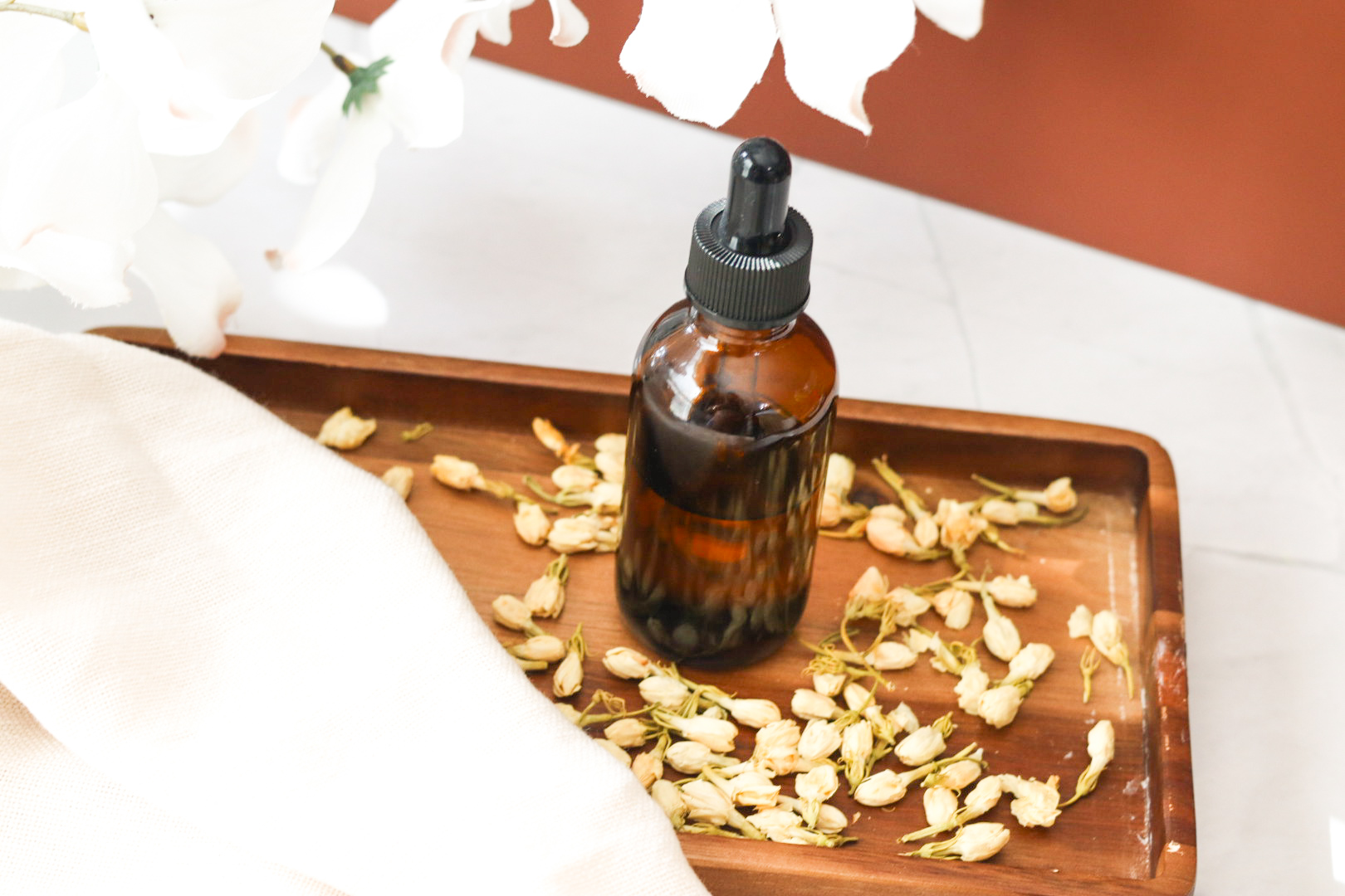 jasmine massage oil in amber dropper bottle next to jasmine leaves and lavender on wooden tray with jasmine buds